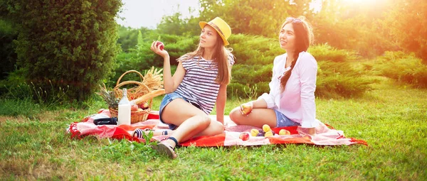 Two beautiful young girls are sitting on a summer picnic in the park at sunset and eating apples Banner concept