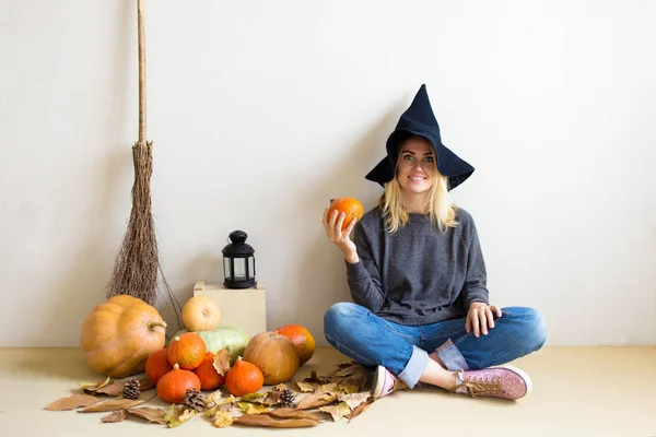 Halloween concept. Beautiful blond girl in a witch hat with pumpkins and broom sitting on the floor against a white wall background Copy space