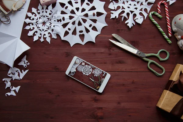 New Year's concept. Smartphone with photo for blog, snowflakes cut from paper, gifts, scissors on a wooden table Copy space