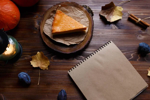 Notebook, pumpkin cheesecake, cooked at home, pumpkin, foliage, table lamp, vanilla on a wooden dark table Copy space