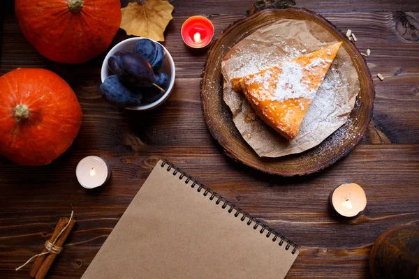 Notebook, pumpkin cheesecake, cooked at home, pumpkin, foliage, table lamp, vanilla on a wooden dark table. Autumn and winter cozy concept.
