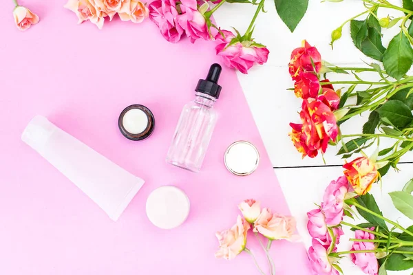 Skin Care Cosmetics in a Frame of Fresh Roses. Essence, Oil on a pink and white desk.