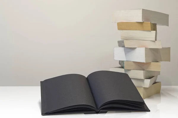 Black Open Book Foreground Pile Books White Table Light Grey — Stock Photo, Image