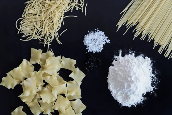 Üç çeşit makarna, un, tuz ve karabiberi yığını ortaya konulan siyah arka plan üzerine