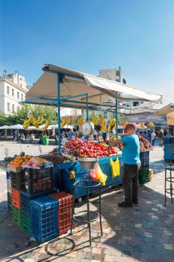 Muz, elma ve mevsimlik sebze satan düzgün düzenlenmiş meyve tezgahlarıyla hareketli bir açık hava pazarı, güneşli bir gökyüzü altında dinamik bir sokak alışveriş ortamı yaratıyor..