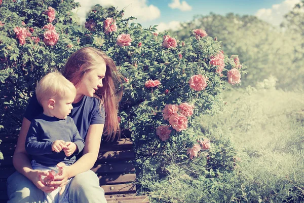 日当たりの良い公園の開花茂みで母親と一緒にベンチに座って幸せな少年 — ストック写真