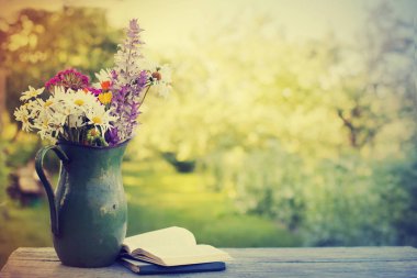 Açık havada kitaplarla ahşap masa üzerinde rustik sürahi içinde renkli taze kır çiçekleri buketi