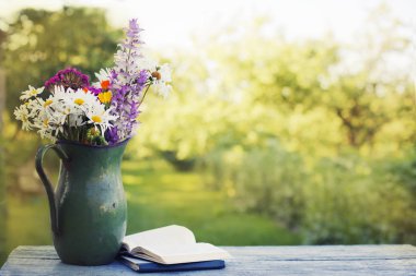 Açık havada kitaplarla ahşap masa üzerinde rustik sürahi içinde renkli taze kır çiçekleri buketi