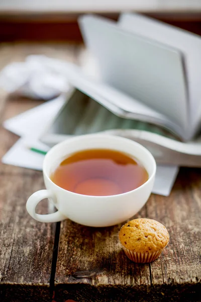 cup of hot tea, muffin and notebooks on wooden surface