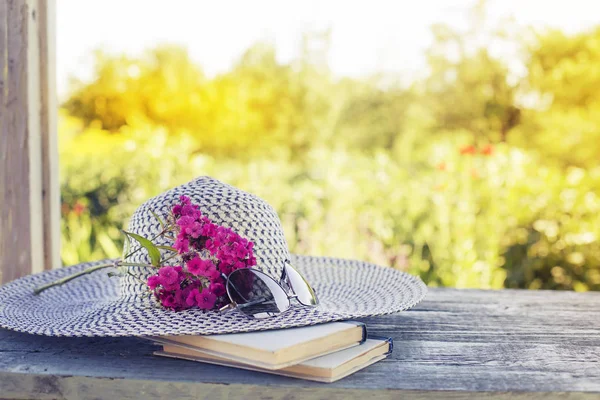 Female hat with glasses, books and pink flowers on blurred nature background