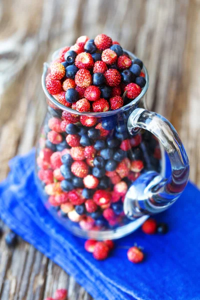 glass pitcher of fresh wild strawberries and blueberries on wooden surface with cloth