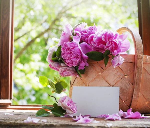 bouquet of fresh cut pink peonies in basket on wooden windowsill with empty card