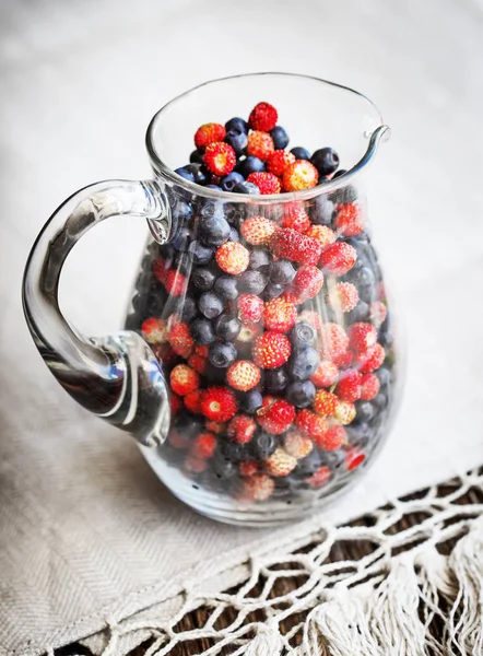 glass pitcher of fresh wild strawberries and blueberries on rural tablecloth