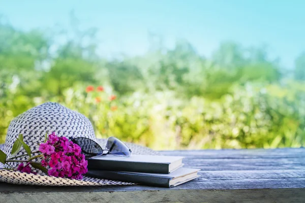 Female hat with glasses, books and pink flowers on blurred nature background