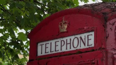 Yıpranmış kırmızı bir İngiliz telefon kulübesi yeşil ağaç yapraklarının altında duruyor, boyası soyuluyor, yakın plan çekim sırasında yakalanmış..