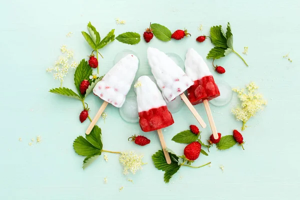 Colorful white and strawberry ice cream on a stick, fruit and berry ice on a blue wooden background with leaves and berries of wild strawberry, top view