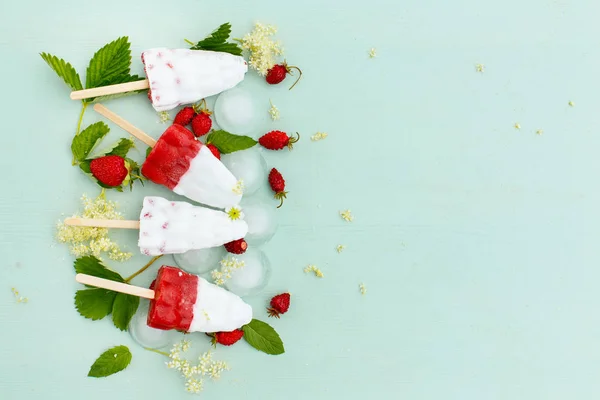 Colorful white and strawberry ice cream on a stick, fruit and berry ice on a blue wooden background with leaves and berries of wild strawberry, top view, space for text