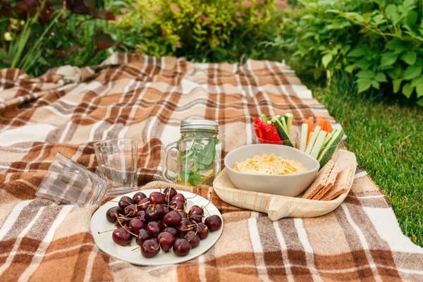 Picnic outside in the Park on the grass wicker box, checkered plaid with food and snacks, hummus and vegetables, cherries, water and glasses
