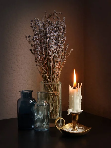 Still life with candlestick with burning candle, essential oil in small vintage bottles and dried bouquet of lavender flowers in the glass vase against low key background. Selective soft focus.