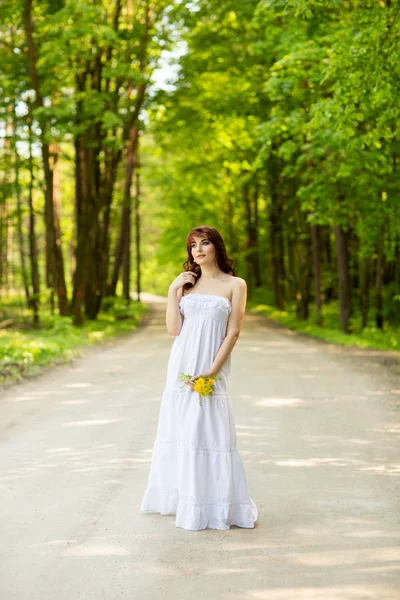 Menina bonita com flores de dente de leão na estrada da floresta — Fotografia de Stock