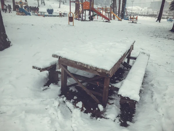 Fából készült asztal és padok a hóban, a fa alatt. Havazás. Gyermekek sétálni a hóban. Hegyi sí resort Bakuriani — Stock Fotó