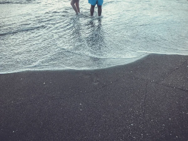 Ноги людей стоят на берегу моря. Блестящие волны на песчаном пляже. Летний солнечный день, песчаный фон — стоковое фото