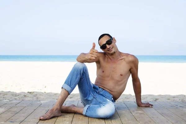 Giovane uomo muscoloso in jeans pantaloni a riposo e in posa sulla spiaggia — Foto Stock