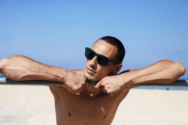 Joven musculoso descansando y posando en la playa. Usar gafas de sol — Foto de Stock