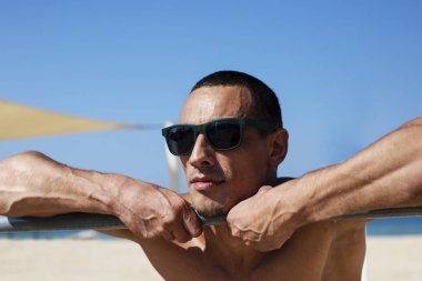 Genç kaslı adam sahilde dinleniyor ve poz veriyor. Güneş gözlüğü takıyor.