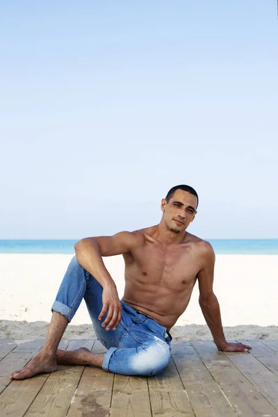 Молодой мускулистый мужчина в джинсовых штанах отдыхает и позирует на пляже — стоковое фото