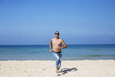 Genç kaslı adam sahilde dinleniyor ve poz veriyor. Bize doğru koş.