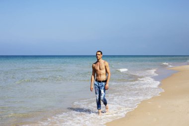 Genç kaslı adam sahilde dinleniyor ve poz veriyor. Genç bir adam deniz kenarında yürüyor.