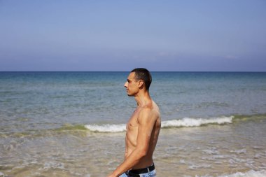Genç kaslı adam sahilde dinleniyor ve poz veriyor. Genç bir adam deniz kenarında yürüyor.