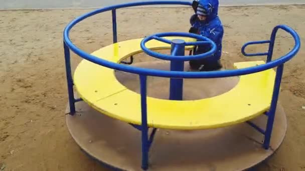 El niño de 4 años en una chaqueta y pantalones se sacude en una rotonda en el patio de recreo — Vídeo de stock