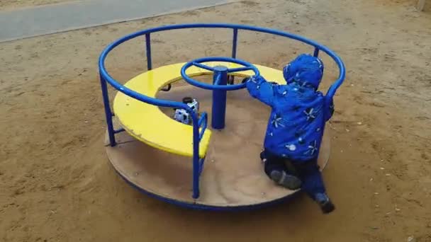 Le petit garçon de 4 ans en veste et pantalon se secoue sur un rond-point dans une aire de jeux — Video