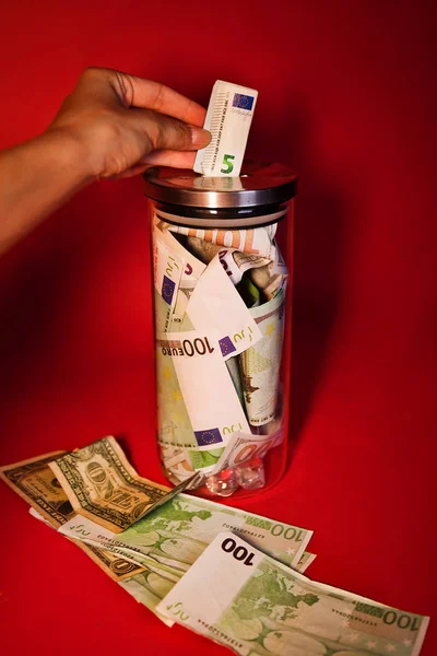 Beautiful Young Woman Hand Drops Euro To The Jar Full Of Money On Red Wall Background