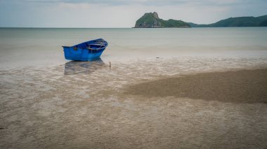 Khao Lummuak 'ın arka planında mavi gökyüzü ve alçak gelgitte kumsalda oturan mavi balıkçı teknesi.
