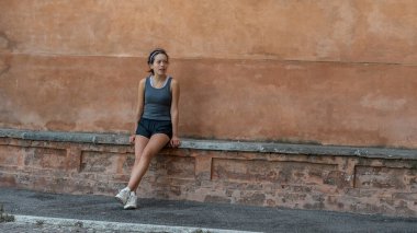 Saç bandı, şort, tanktop ve spor ayakkabılı genç kız Bolgona İtalya 'da tuğla bankta oturuyor.