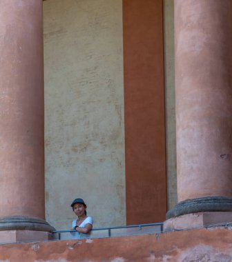 Bologna İtalya 'da büyük sütunlar arasında balkonda duran tişörtlü ve şapkalı Asyalı kadın.