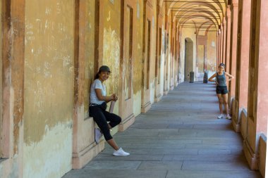Şapkalı anne elinde termosla duvara yaslanırken genç kız Bolonya İtalya 'da kalçasında elleriyle ona bakıyor.