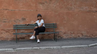 Şapkalı, tişörtlü, siyah pantolonlu ve spor ayakkabılı bir kadın Bologna 'da bacak bacak bacak üstüne atmış bir şekilde park bankında oturuyor.