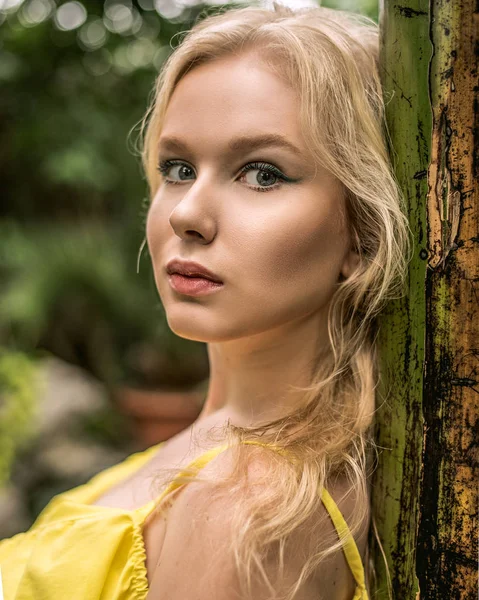 En vacker ung garvade flicka med huvudduk på hennes huvud och bära solglasögon står nära en gul vägg i söder. Fashionabla blå sommarklänning, ljusa make-up, tan. Palms, skugga. — Stockfoto