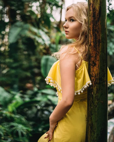 Ein schönes, braungebranntes Mädchen mit Kopftuch und Sonnenbrille steht an einer gelben Wand im Süden. modisches blaues Sommerkleid, helles Make-up, Bräune. Palmen, Schatten. — Stockfoto