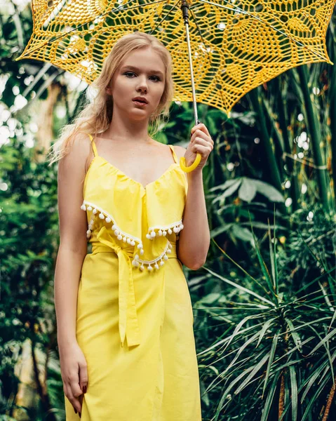 En vacker ung garvade flicka med huvudduk på hennes huvud och bära solglasögon står nära en gul vägg i söder. Fashionabla blå sommarklänning, ljusa make-up, tan. Palms, skugga. — Stockfoto