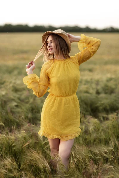 Menina sonhadora loira em um campo de trigo usando um vestido amarelo brilhante e um chapéu de palha . — Fotografia de Stock