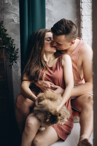 Beau jeune couple blanc européen avec un chat sur les mains assis sur un canapé gris à la maison par une journée ensoleillée. Embrasser, heureux, pull, isolement, quarantaine, coronavirus, animal de compagnie. — Photo