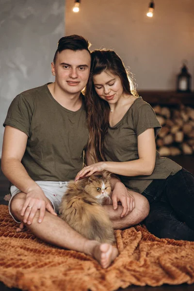 Vackra unga europeiska vita par med en katt på händerna sitter på en grå soffa hemma på en solig dag. Omfamna, glad, tröja, isolering, karantän, coronavirus, husdjur. — Stockfoto
