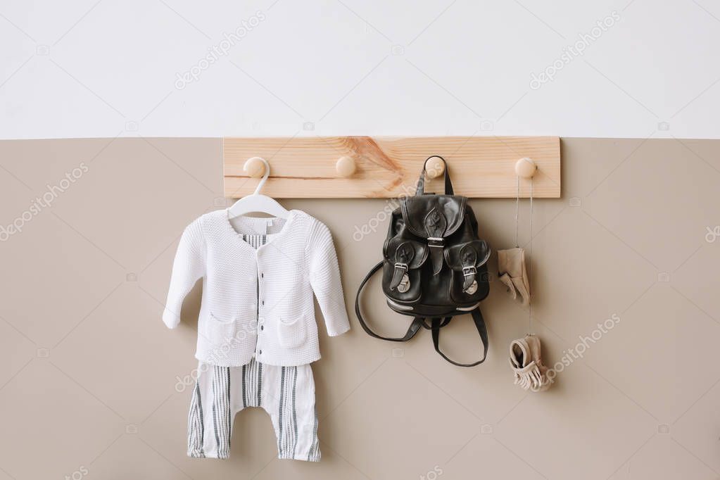 Interior details of children's room on light background. Decorative wooden hanger with bag and children's knitted costume on white and brown wall 