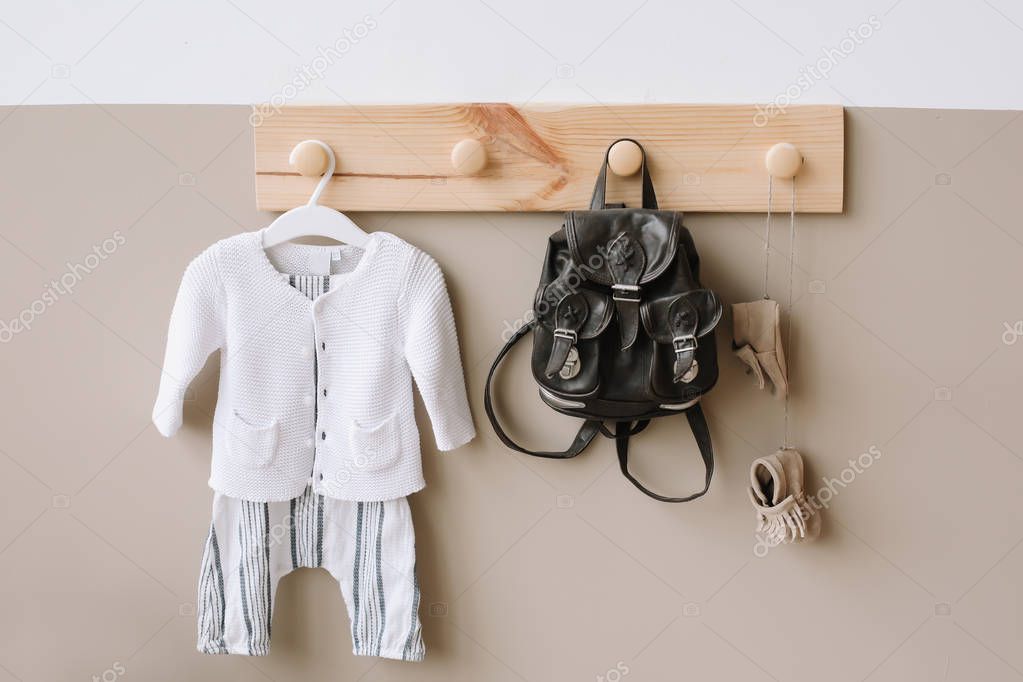 Interior details of children's room on light background. Decorative wooden hanger with bag and children's knitted costume on white and brown wall 