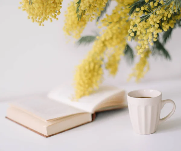 Xícara de café e um livro sobre mesa branca com flores amarelas bouquet mimosa. Conceito de planejamento e design. Deitado. Vista superior . — Fotografia de Stock